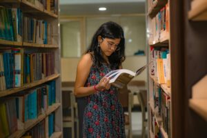 A South Asian woman with glasses reads attentively in a Delhi library, surrounded by bookshelves.