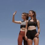 Two fit young women in workout attire posing confidently against a clear blue sky.