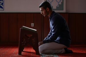 A man praying inside a mosque, kneeling beside a Quran on a stand.