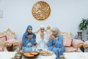 Three Muslim women in hijabs pray indoors during Ramadan, sitting on an ornate couch with traditional food.